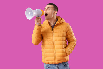 Young man in warm puffer jacket shouting into megaphone on purple background