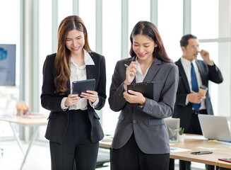 Asian professional successful female businesswomen in formal suit standing smiling together using touchscreen tablet computer surfing browsing internet and writing note on paper clipboard in office