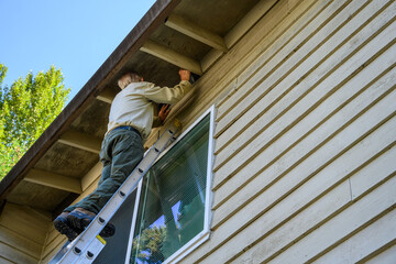 Senior man on an extension ladder replacing the mesh of an attic vent on the exterior of a...