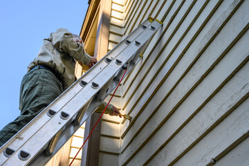Senior man on an extension ladder painting sealer on wood siding of the exterior of a residential building
