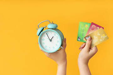 Female hands holding alarm clock and credit cards on yellow background