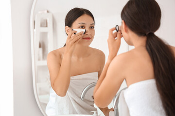 Young Asian woman removing makeup with reusable cotton pad near mirror in bathroom