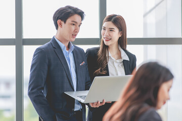 Asian professional successful female businesswoman and male businessman colleague employee in formal suit standing smiling together browsing surfing internet online via laptop and tablet in office