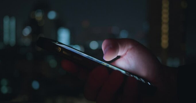 Close up person use mobile phone at night time on urban light background. Cinematic film look of male hand use mobile phone, tapping and scrolling news feed on app on his phone at night