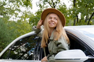 Blonde woman in hat sticking head out of windshield car. Young tourist explore local travel making...