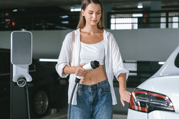 Young woman travel with EV electric car to shopping center parking lot charging in downtown city...