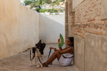 A brunette woman, seated on a street, rests and refreshes herself with a fan, accompanied by her dog