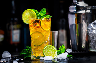 Dark and Stormy cocktail drink with dark rum, ginger ale, lime and ice with bottles, black bar...