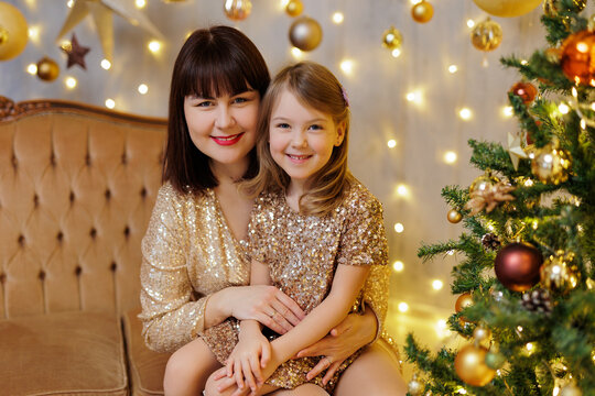 Mother and little daughter in golden dresses sitting on sofa in christmas decorated room