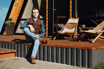 Middle-aged man in jeans, shirt and vest sitting on terrace of wooden cabin