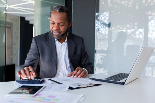 Successful Satisfied Financier Behind Paper Work, Afro American Senior Experienced Filling Out Documents, Using Calculator For Calculation, Businessman Behind Paper Work Inside Office Smiling.