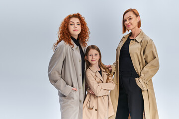 three generation joyful family standing together in stylish coats on grey backdrop, autumn fashion