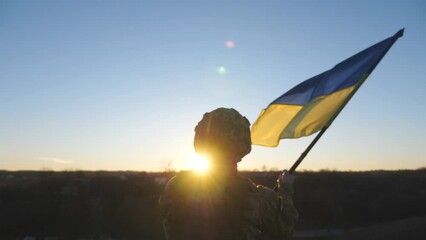 Young man in military uniform waving flag of Ukraine against background of sunset. Male ukrainian army soldier lifting national banner in hill. 