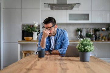 Exhausted and anxious businessman with head in hand sitting by laptop at desk in home office