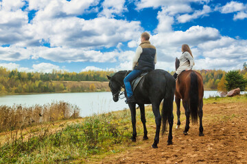 Rear view of couple man and woman on horses in fall forest with lake by blue sky. From behind...