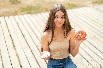 Teenager girl holding a donut at outdoors making doubts gesture while lifting the shoulders