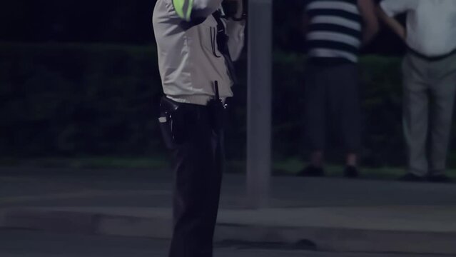 Highway patrol officer guard with handheld light stick baton directs traffic at night. Pedestrians cross roadway in background