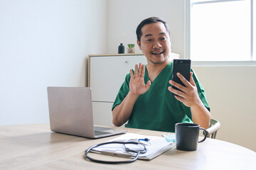 Cheerful young Asian male doctor waving hand say hi at mobile phone during tele medicine
