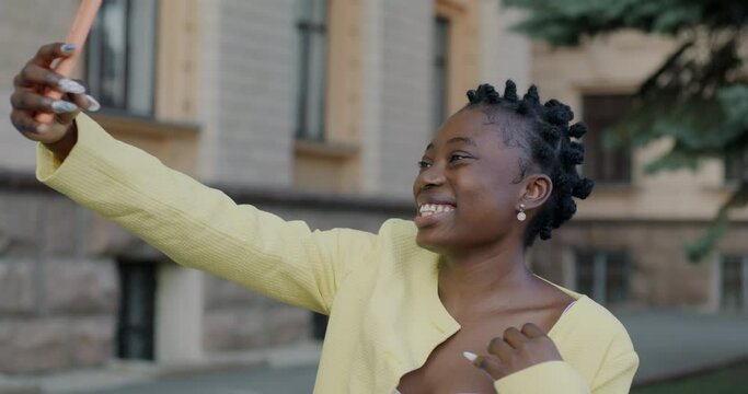 Slow motion portrait of pretty African American girl taking selfie posing for smartphone camera in urban street. Photography and gadgets concept.
