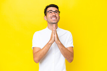 Young handsome man over isolated yellow background keeps palm together. Person asks for something