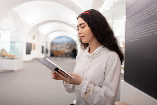 Side View Of Young Caucasian Woman Student Visiting Museum Exhibition And Reading Brochure. Light Hall In Background. Concept Of Cultural Education And Museum's Day