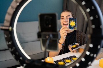 Young woman fortune teller recording video for blog using tarot cards, smartphone and ring lamp, predicts future online from home office