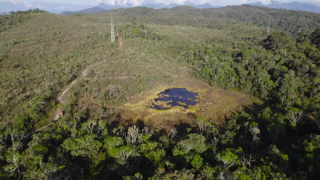 Cinematic daytime drone footage in Oxapampa, Peru. The camera ascends slowly while gradually tilting downward, revealing green-covered lake nestled among the forest with power lines in the background.