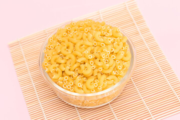 Uncooked Chifferi Rigati Pasta in Glass Jar on Bamboo Mat on Pink Background. Fat and Unhealthy Food. Classic Dry Macaroni. Italian Culture and Cuisine. Raw Pasta
