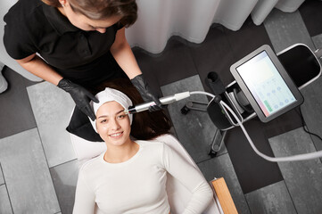 Top view of smiling woman lying near cosmetology equipment and receiving facial treatment....