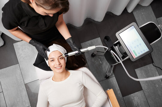 Top view of smiling woman lying near cosmetology equipment and receiving facial treatment. Cosmetologist using radiofrequency microneedling device while performing beauty skincare procedure.
