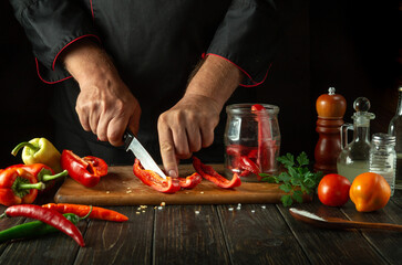 Slicing sweet red pepper on wooden cutting board. Professional cook preparing salad. Male hand cuts capsicum with knife. Restaurant staff at work