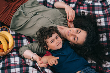 Top view on laying on plaid curly young woman with little son, having picnic outdoors. Little child points at camera, spends time with mom. Motherhood, leisure, family moments.