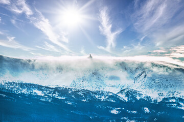 Zdjęcie przedstawia górski szczyt spowity gęstą mgłą spływającą po zboczu. Wśród mgły widać zarys budynku. Niebo jest lekko zachmurzone, świeci słońce. - obrazy, fototapety, plakaty