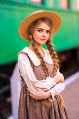 Young pretty blonde braided girl in vintage dress and straw hat standing on station platform near green train. Portrait of Smiling elegance cute lady traveller with blue eyes .
