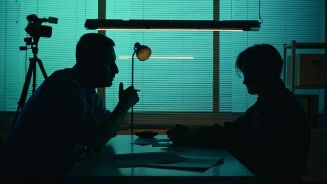 Medium silhouette video of a offender, perpetrator or prisoner sitting in the interrogation room in front of the detective. Suspect tries to strangle the policeman, but gets stopped.