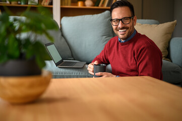 Portrait of businessman smiling and holding coffee cup while working over laptop on sofa at home