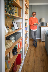 Full length of smiling young businessman reading book while leaning on bookshelf in home office