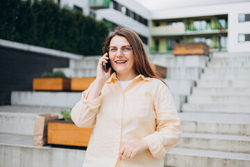 Lifestyle portrait of a stylish young 30s businesswoman talking on cell phone, making call and standing on modern city street. Successful small business. Urban lifestyle concept.