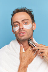 Close-up of handsome man with under eyes patches and shaver in his hands, morning procedures,...