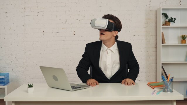 Medium shot of a young man in an office wearing virtual reality, vr helmet, gear, looking at, interacting with an invisible screen excited and happy.