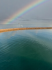 Two seagulls fly over a harbor pier. In the background a rainbow over the sea.