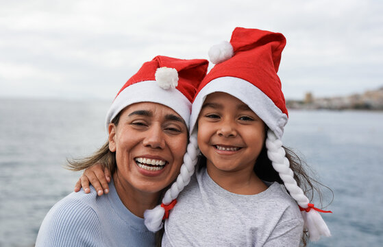 Happy latin mother and little daughter wearing christmas hat with beach in background