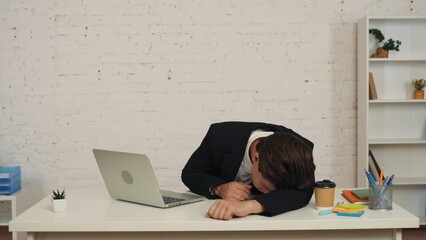 Medium shot of a young man sitting at the table in front of the laptop in the office, laying on his...