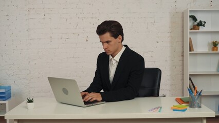 Medium shot of a young businessman sitting at the table in an office, working on his laptop, focused, consentrated and seious.