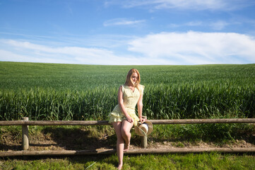Blonde, young, pretty farm woman, sitting on a wooden fence. In the background a green wheat field....