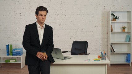 Medium shot of a young man leaning, standing next to his working space, table, holding a coffee cup and looking to the side pensively.