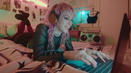 Pink-haired girl with headphones resting on bed in 90s aesthetic room with neon light, typing message on laptop and smiling during night at home. Whip zoom shot