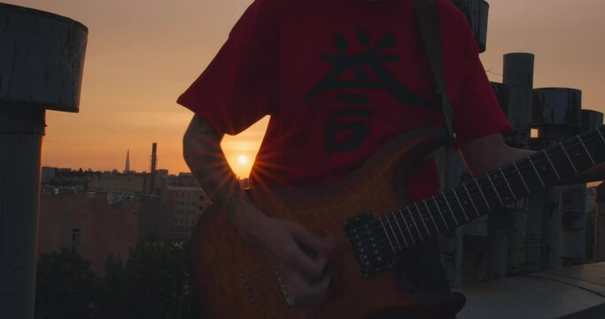 Young tattooed female rocker expressively plays an electric guitar with a pick at dusk at sunset, close up to musical instrument, front view. Outdoor jam session or rehearsal.