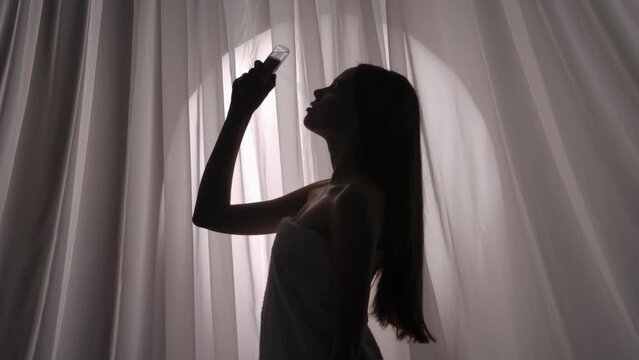 Medium side view video of a young woman silhouette wrapped in a towel using a facial mist sprayer, moisturizing device on her skin in a muffled light.