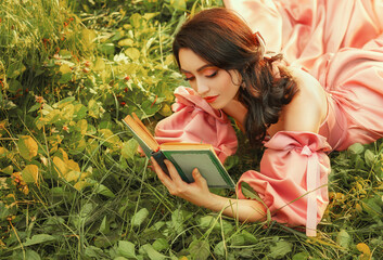 Fantasy happy woman lies on green grass of forest lawn holds in hands magic book novel old style, bright magical sun light. Romantic cute sexy girl in long pink vintage dress. Medieval lady magician